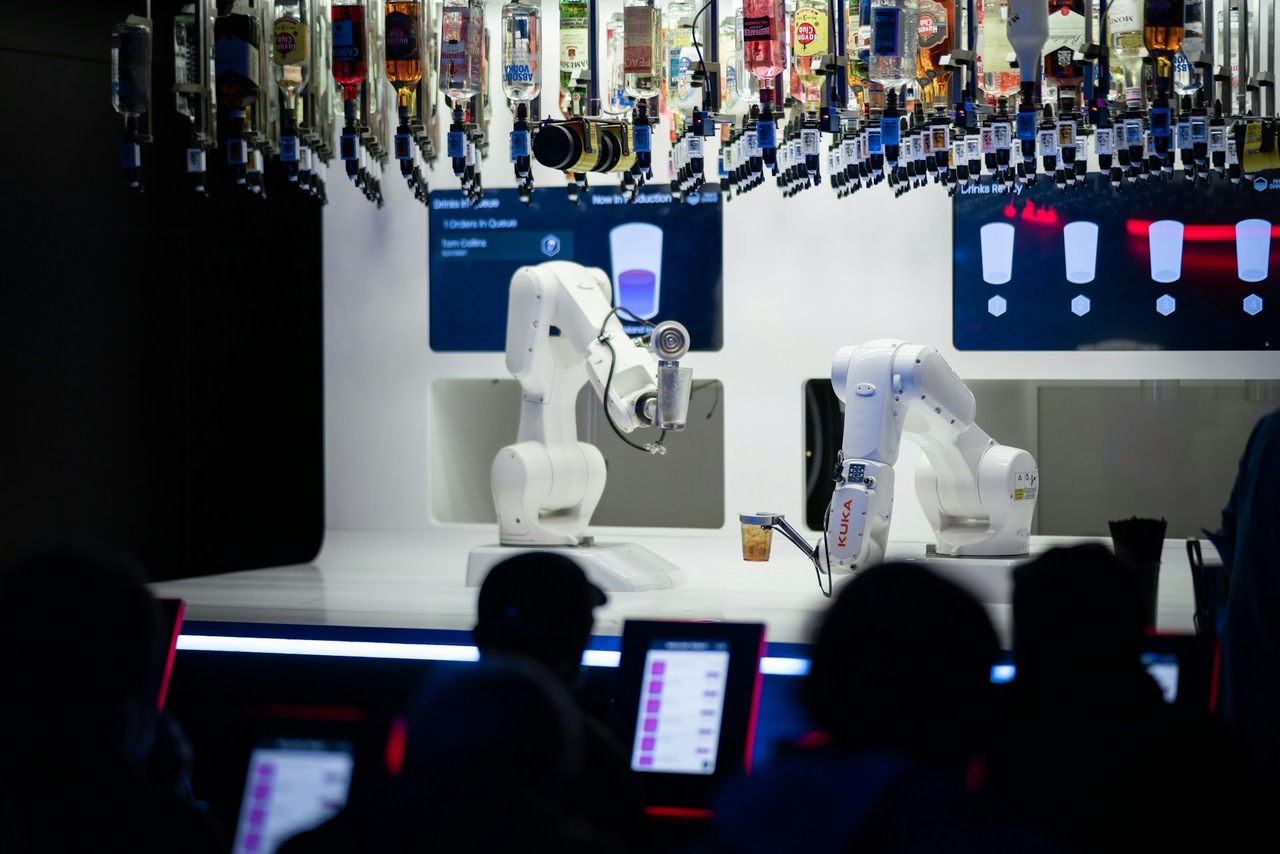 a group of people watching how two robotic arms perform bartender tasks